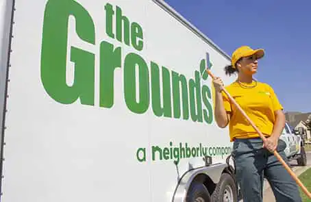 Grounds Guys worker in yellow polo and hat, holding rake, standing next to trailer displaying company logo.
