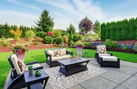 A backyard patio with lush green lawn and beautiful landscaping behind it.