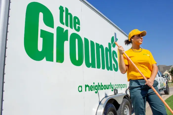 Grounds Guys employee wearing yellow hat and shirt, holding rake and standing next to branded company trailer.