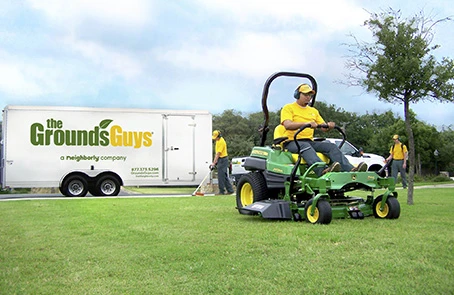 Grounds Guys employee in branded shirt using riding mower to cut grass, with company trailer in background.