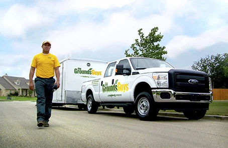 Grounds Guys associate in yellow T-shirt beside branded pickup truck and trailer.