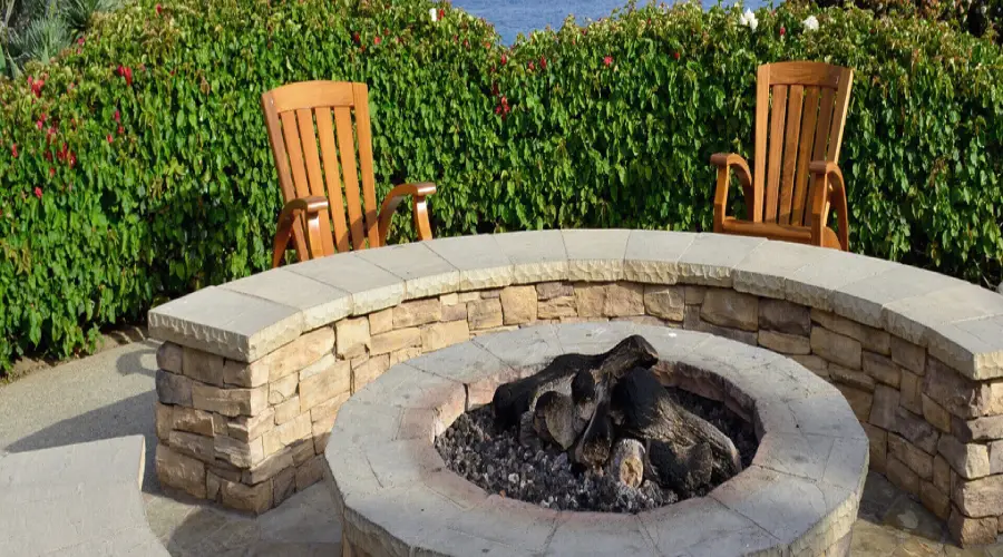 Stone firepit with wraparound stone bench, Adirondack chairs, and hedge.