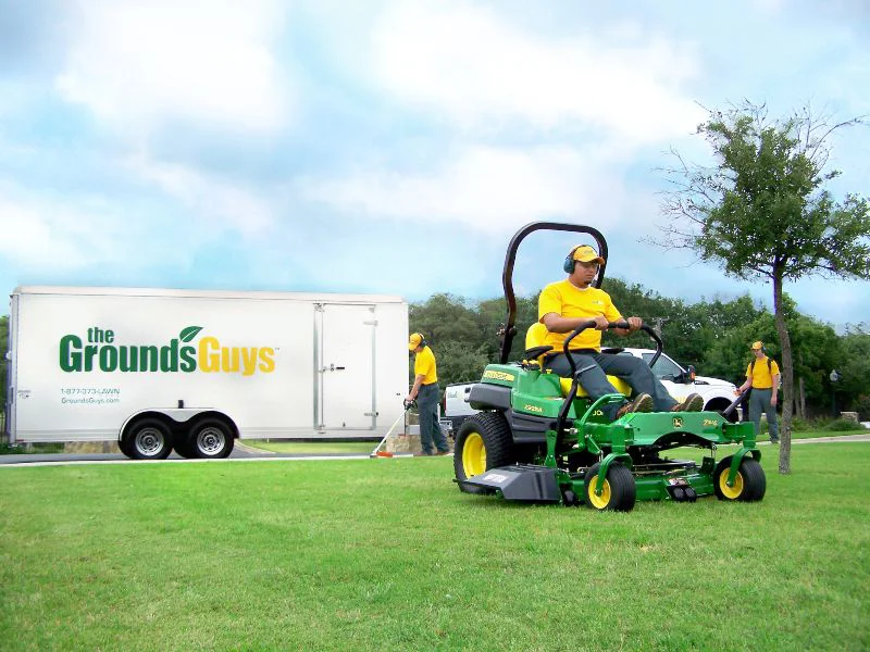 The Grounds Guys employee mowing a bright green lawn with a riding lawnmower.